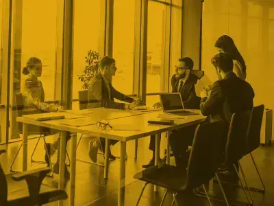 Professionals in a modern meeting room discussing strategy around a conference table with laptops and documents.